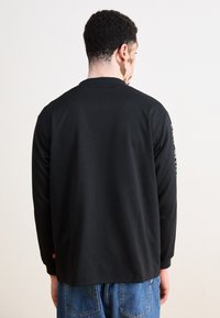 Schwarzes Langarmshirt mit Rundhalsausschnitt, mit einem grünen und weißen Logodetail am linken Ärmel. Glattes Baumwollmaterial, lockere Passform.