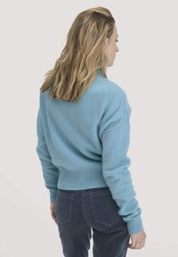 Frau mit schulterlangem blonden Haar, die einen hellblauen Pullover und eine dunkelblaue Cordhose trägt, von hinten vor einem einfarbigen Hintergrund.