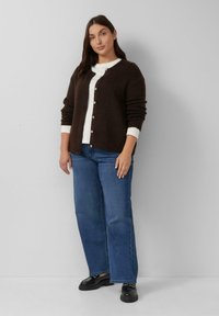 Brauner gestrickter Cardigan mit Knopfleiste, weißen Bündchen, kombiniert mit blauen Jeans im geraden Schnitt und glänzenden schwarzen Schuhen. Einfaches Design.