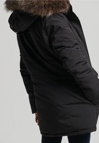 Černá prošívaná parka s kapucí lemovanou kožešinou, s hladkou texturou, dlouhými rukávy a střední délkou pro teplo a ochranu.