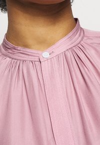 Rosa blus med plisserad design, med mandarin-krage och vit knäppning, tillverkad av slät tyg.