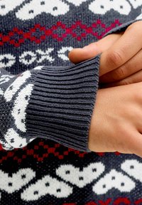 Fonott pulóver sötét szürke bordás mandzsettával, fehér szívecskék és piros kiegészítők mintájával, sötét háttéren.