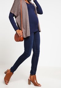 Těhotenský outfit složený z námořnického topu, pleteného taupe kardiganu, tmavých skinny džínů a hnědých sandálů na podpatku s otevřenou špičkou, doplněný hnědou kabelkou na přes rameno.