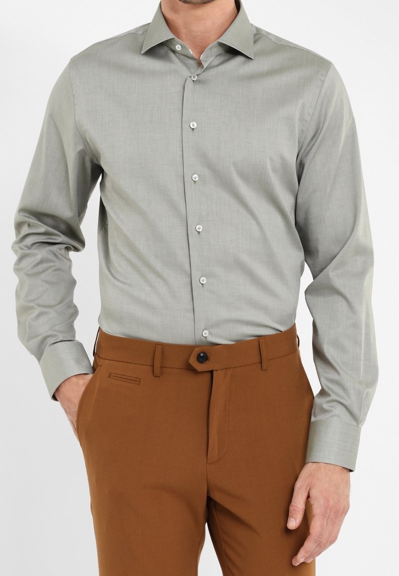 Homme portant une chemise à manches longues gris clair boutonnée, rentrée dans un pantalon marron, une main dans la poche, sur fond blanc uni.