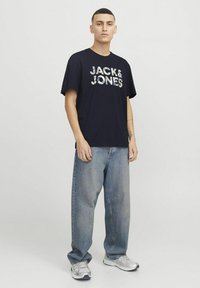 Tamsiai mėlynas marškinėliai su balta "JACK & JONES" užrašu, derinami su šviesiai mėlynomis plačiomis džinsomis ir pilkomis sportinėmis bateliais, stovint ant vienspalvio fono.