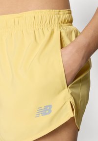 Gula sportshorts med resårmidja, sidofickor och en subtil New Balance-logotyp i grått på nedre vänstra sidan.