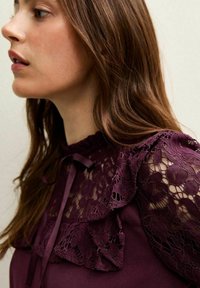 Vestido burdeos con mangas de volantes de encaje, cuello con lazo y tela suave, con patrones detallados de encaje y textura en las mangas.