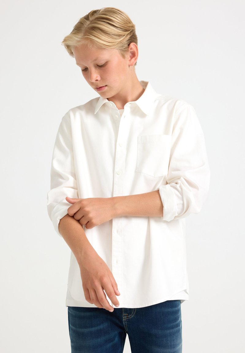 Hvid langærmet skjorte med knappet krave, enkelt brystlomme og op-rullede ærmer, kombineret med mørkeblå jeans.