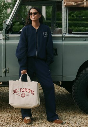 Tumesinine lukuga dressipluus ja sobivad püksid pehmes kangas. Käes beežikas nailonist kott punase "SELFSports" tekstiga. Pruunid seemisnahast kingad.