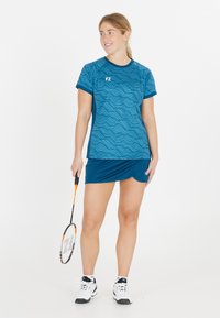 Blå atletisk skjorte med bølgemønster, matchende blå nederdel og hvide sneakers. Modellen holder et badmintonracket og smiler.