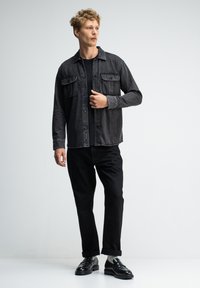 Zwarte denimoverhemd met twee borstzakken, gedragen over een zwart t-shirt. Zwarte broek en glanzende zwarte schoenen maken de outfit compleet.