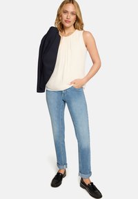 Blouse blanche sans manches à plis, jean bleu retroussé, et mocassins noirs ; blazer noir posé sur un bras.
