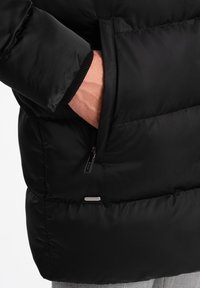 Giacca a vento nera con superficie testurizzata, dotata di una tasca con zip, polsini a coste e una forma arrotondata. I dettagli includono un'etichetta con logo.