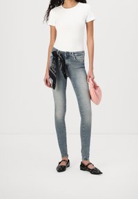 Vaqueros ajustados en denim azul deslavado, camiseta blanca ajustada, bandana negra anudada a la cintura y zapatos negros con recortes circulares. Sosteniendo una bolsa rosa.