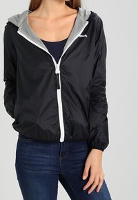 Černá bunda na zip vyrobená z lehkého materiálu, s šedou kapucí s podšívkou z síťoviny, elastickými manžetami a bílým zipem.