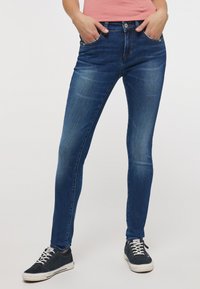 Jeans skinny azul oscuro con bolsillos frontales, áreas desgastadas y cierre de botón. Combinados con zapatillas marinas que tienen cordones y suelas blancas.