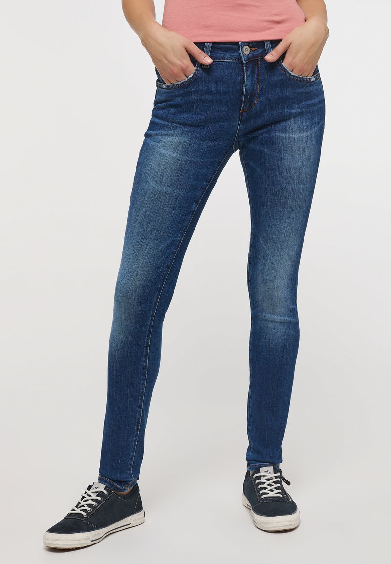 Jeans skinny azul oscuro con bolsillos frontales, áreas desgastadas y cierre de botón. Combinados con zapatillas marinas que tienen cordones y suelas blancas.