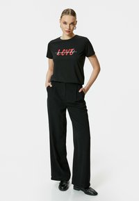 Camiseta negra con gráfico rojo de "LOVE", combinada con pantalones negros holgados. El modelo posa con las manos en los bolsillos, llevando zapatos negros. Fondo blanco liso.