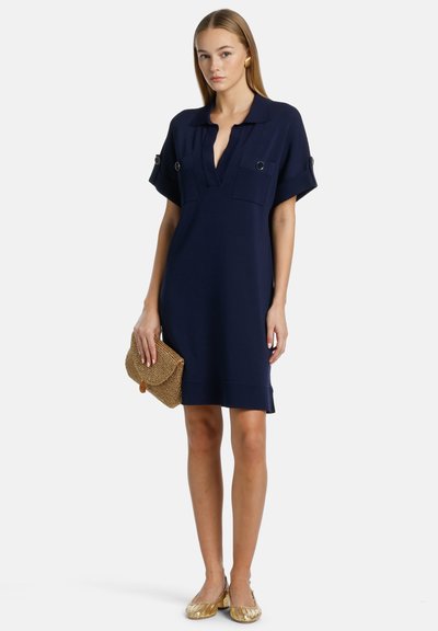Abito blu navy con colletto, maniche corte e due tasche frontali. Tessuto liscio con vestibilità comoda, abbinato a una clutch in paglia e a ballerine dorate.