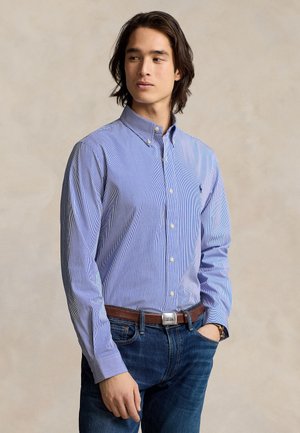 Νέος άνδρας που φοράει μπλε ριγέ πουκάμισο με κουμπιά, φορεμένο μέσα σε μπλε τζιν με καφέ ζώνη, στέκεται μπροστά σε μια απλή φόντο.