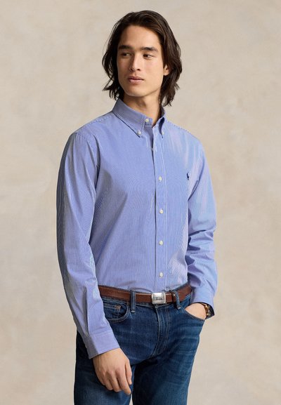 Jeune homme portant une chemise à rayures bleues boutonnée, rentrée dans un jean bleu avec une ceinture marron, debout devant un fond uni.