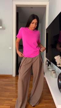 Femme ajustant son t-shirt rose vif rentré dans un pantalon ample beige à jambes larges, debout dans une pièce moderne avec un sol en bois.