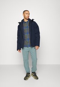 Chaqueta azul marino con capucha, camisa a cuadros verde y azul, pantalones de chándal azul claro, y zapatillas verdosas con suelas negras.