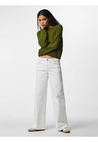 Jeune femme aux cheveux bouclés portant un pull vert, un pantalon blanc à jambes larges et des chaussures blanches, debout avec la main sur le visage.