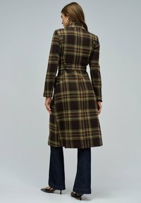 Abrigo de cuadros marrón con cintura ceñida por un cinturón, mangas largas y cuello de muesca. Presenta acentos verdes y rojos en el patrón a cuadros.