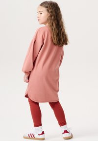 Helles rosa Kleid mit langen Ärmeln und entspannter Passform, kombiniert mit rostfarbenen Leggings und weißen Schuhen mit roten Akzenten und braunen Sohlen.