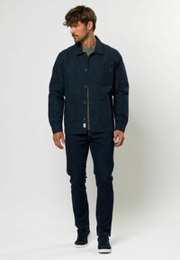 Chaqueta azul marino con cierre de botón, dos bolsillos frontales y un bolsillo en el pecho. Llevada sobre una camisa verde y combinada con jeans oscuros y zapatillas.