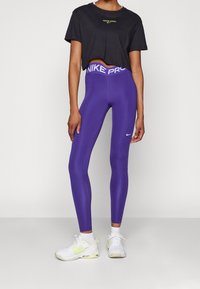 Sort cropped t-shirt med "NIKE PRO" logo, lilla leggings med høj talje og hvide sneakers med gule detaljer. Glat tekstur.