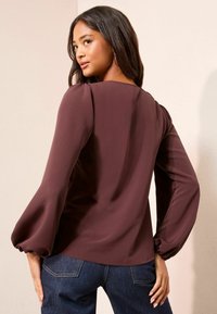 Tumši burgundijas blūze ar pūkainām garajām piedurknēm un apaļu kakla izgriezumu, izgatavota no gludas auduma, apvienota ar tumšām džinsu biksēm.