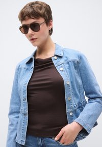 Ανοιχτό μπλε τζιν μπουφάν με ασημένια κουμπιά και εφαρμοστή γραμμή, φορεμένο πάνω από καφέ μπλουζάκι. Το μοντέλο φοράει επίσης σκουρόχρωμα γυαλιά ηλίου.