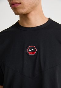 Melns kokvilnas T-krekls ar sešstūra sarkanu un melnu plāksteri ar Nike logo, izgatavots no gludas auduma ar vienkāršu dizainu un bezšuvju kaklu.