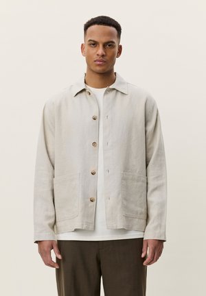 Junger Mann mit kurzen Haaren, trägt eine beige Jacke mit Knöpfen über einem weißen Hemd und dunkelbraune Hose, stehend vor einem schlichten hellen Hintergrund.