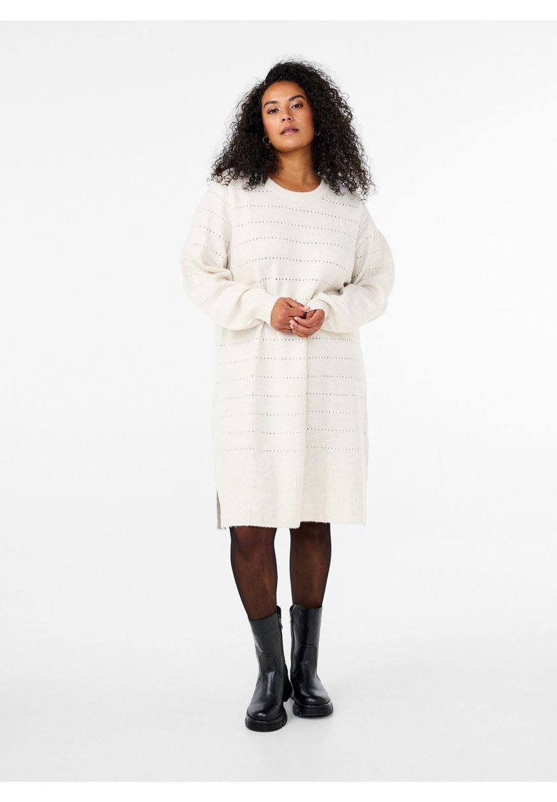 Femme aux cheveux bouclés portant une longue robe-pull blanche, des collants noirs et des bottines noires, debout devant un fond blanc uni.