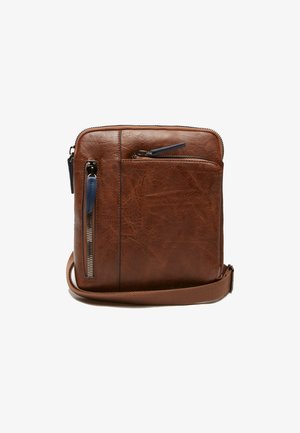 Rudos odos per petį nešiojama rankinė su tekstūruotu paviršiumi, turinti priekinį užtrauktuką, reguliuojamą dirželį ir kontrastingus metalinius akcentus.