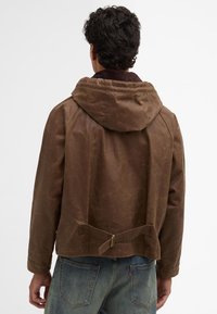 Brown waxed cotton jacket with a hood, featuring a belted waist detail and textured surface, paired with blue denim jeans.