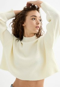 Suéter cortado color crema con textura acanalada, mangas anchas y cuello alto. Material suave con un corte holgado y dobladillo sin acabado.