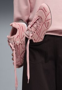 Person trägt pinkes Sweatshirt und hält pinke Turnschuhe mit roten Linien und Texten, die Fleischstücke darstellen, vor grauem Hintergrund.