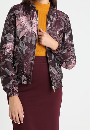 Frau trägt eine lila Blumen-Reißverschlussjacke über einem senffarbenen, gerippten Oberteil und einem bordeauxfarbenen Rock, mit einer Hand nahe am Reißverschluss.