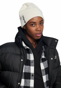 Gorro crema con logo, chaqueta de plumas negra y camisa de cuadros debajo. Presenta trenzas oscuras y un fondo neutro.