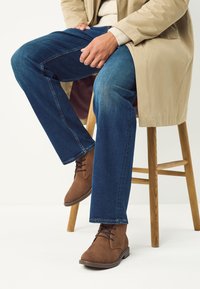 Braune Wildleder-Stiefeletten mit Schnürsenkeln, blaue Denim-Jeans und ein beiger Mantel. Glatte Texturen, schlichtes Design und klassische Passform.