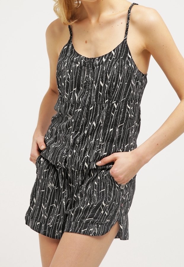 Femme portant un haut sans manches noir à boutons et un short assorti avec motif de plumes blanches, mains dans les poches, debout devant un fond uni.