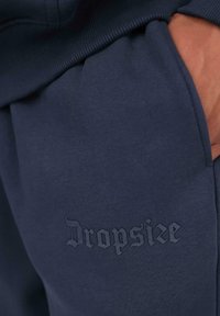 Marineblaue Sweatpants mit eingeprägtem "Dropsize"-Logo in der Nähe der Tasche, Hand teilweise in der Tasche.