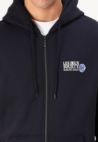 Marineblaue Zip-Hoodie mit Kängurutasche, Zugkordelkapuze und weißem Logo-Print auf der linken Brust. Hergestellt aus weichem Baumwollmischgewebe.