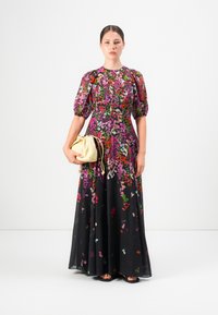 Vrouw in een lange, zwarte bloemenjurk met pofmouwen, met een kleine beige trekkoordhandtas, staand tegen een witte achtergrond.