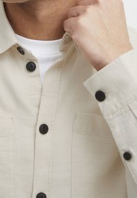 Beige Hemd mit Knöpfen in Schwarz, ausgestattet mit zwei Brusttaschen und einem strukturierten Stoff. Sichtbare weiße Innenschicht am Kragen.