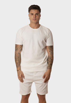 Camiseta blanca de textura con mangas cortas, cuello redondo y un sutil detalle de logo, combinada con pantalones cortos a juego. En general, un conjunto coordinado.